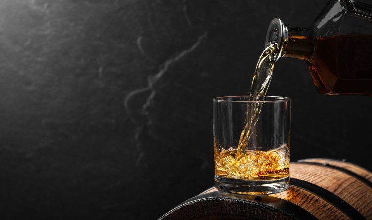 Pouring whiskey into glass from bottle on wooden barrel against grey background