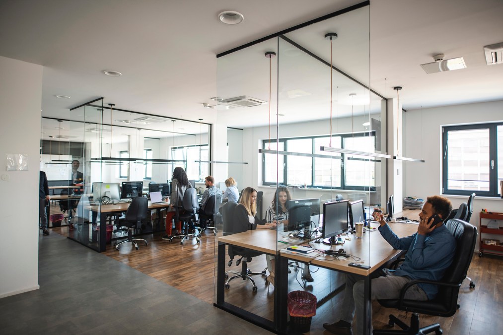 Young adult business colleagues interacting at communal workstations in see-through open plan office space.